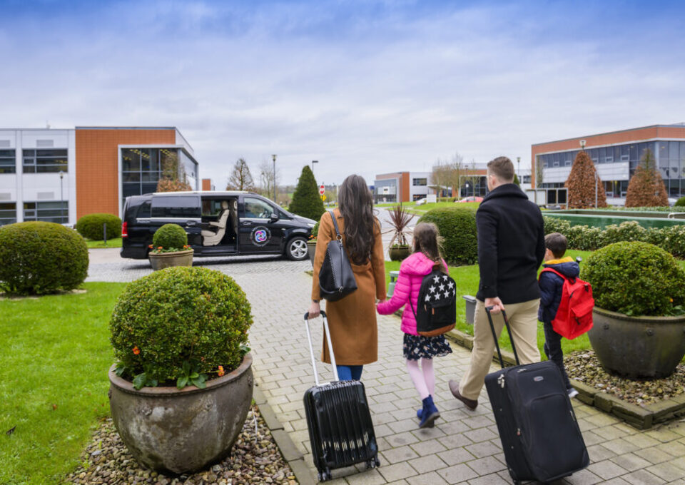 Family with suitcases walking out the front of the hotel to the Hotel Shuttle bus with smiling driver